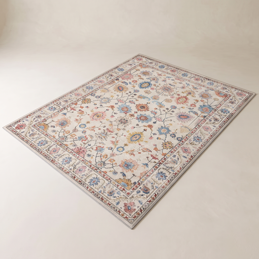 Bahia Bohemian Velvet Rug shown flat on a pale plaster floor, ivory ground with a faded pastel Persian floral repeat and a wide brick-rust bordered frame