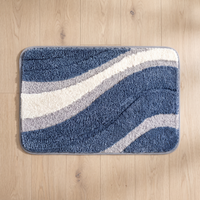 Nima bath mat seen from above on a pale oak floor, showing the full 50 x 80 cm rectangle, its sweeping indigo, ivory and dove grey arcs, rounded corners and grey bound edge