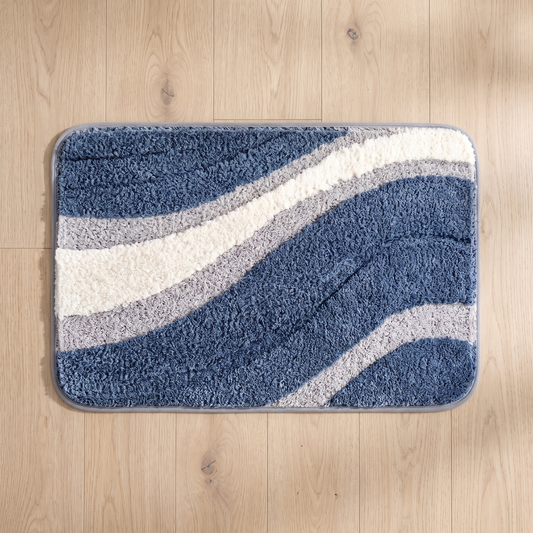 Nima bath mat seen from above on a pale oak floor, showing the full 50 x 80 cm rectangle, its sweeping indigo, ivory and dove grey arcs, rounded corners and grey bound edge