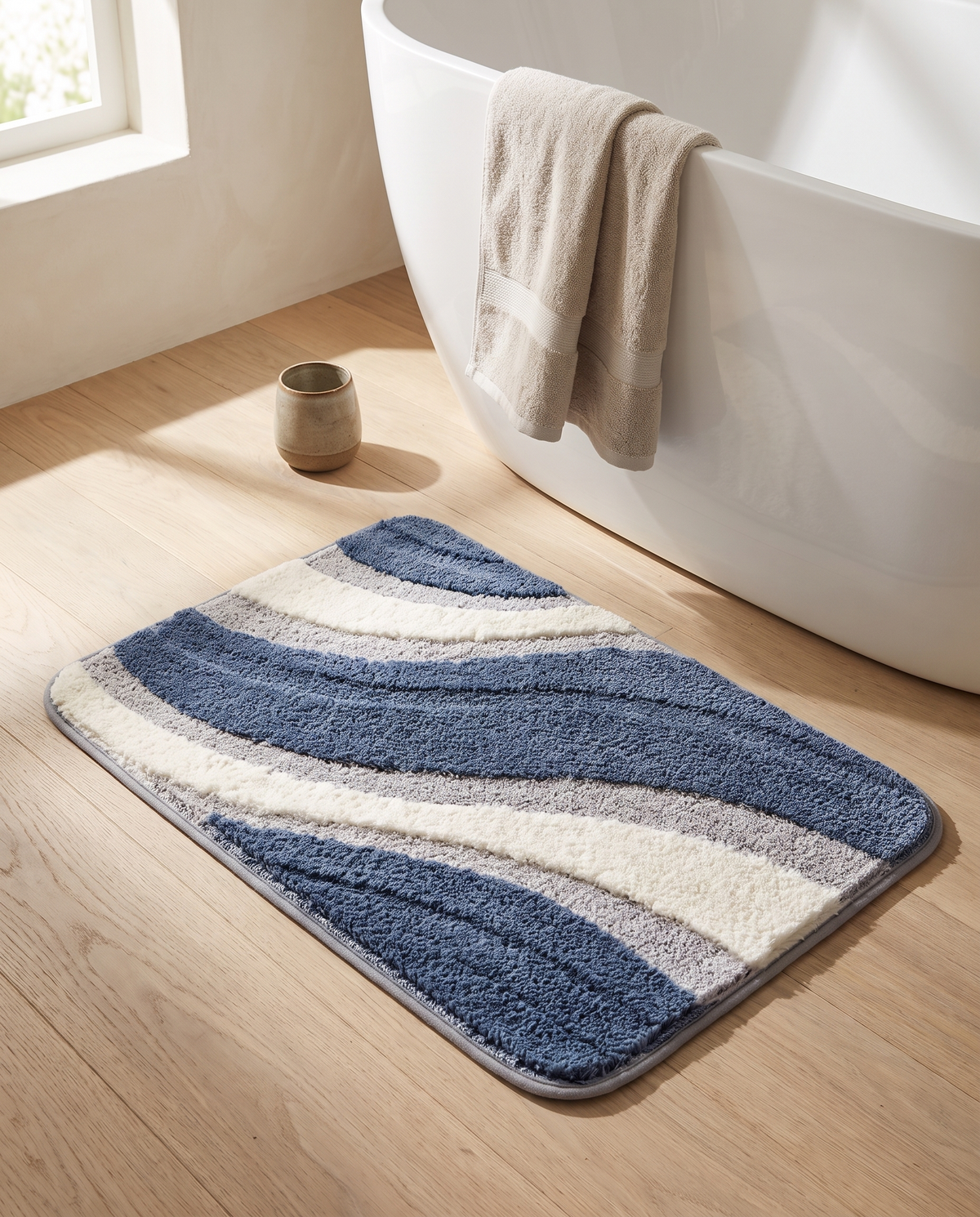 Nima bath mat on a pale oak floor beside a freestanding white bath in a warm neutral UK bathroom, with a folded linen towel over the rim