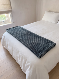 Nordo grey throw blanket folded lengthwise across the foot of a bed dressed in white linen, showing its lap-throw scale