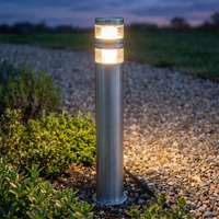 Vestro path post light glowing in a planted border beside a gravel garden path at dusk