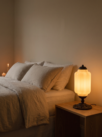 Elva Glass Table Lamp - lit warm on oak bedside table in linen bedroom, glowing 2700K amber