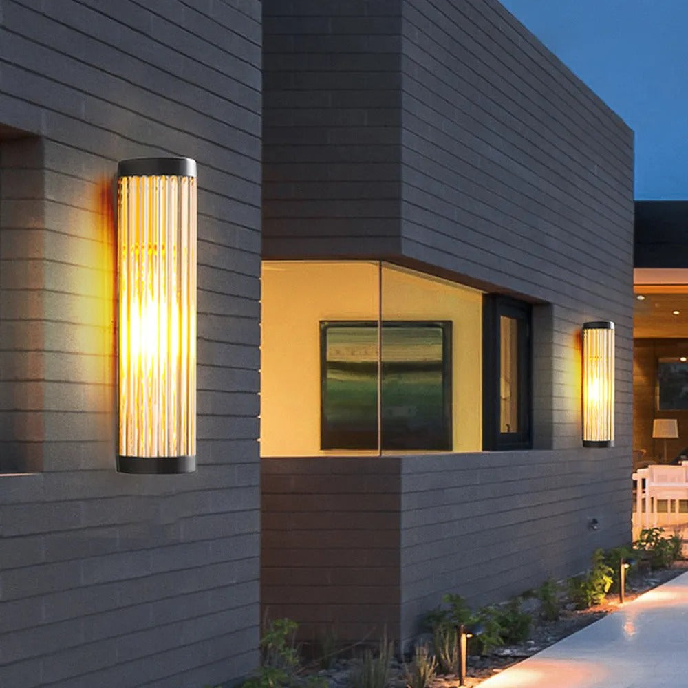 Yoro wall light lit on a grey brick house exterior at twilight, casting warm light up and down the wall